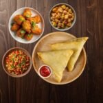 Crispy samosa platter with chutney, fried appetizers, and chickpea snack served on wooden table