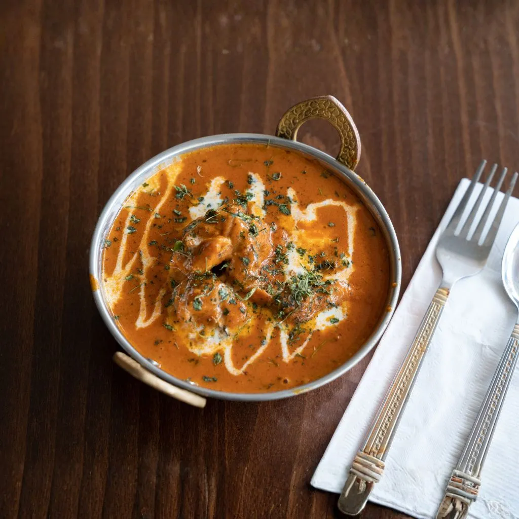 A metal bowl of creamy butter chicken topped with herbs and drizzled cream, served on a wooden table with cutlery on the side, showcasing a rich and comforting tomato based curry.
